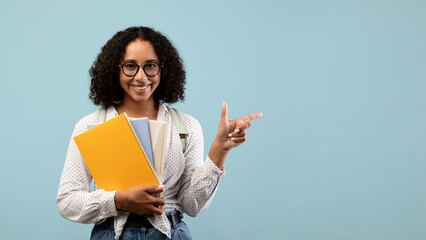 Obraz premium A joyful black female student with a backpack holds study materials and points aside at an empty space for your advertisement. She is ready to share educational offers on a light blue background.