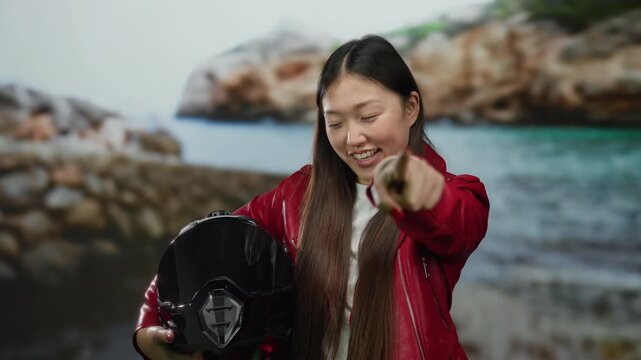 Woman smiling in red jacket holding helmet at seaside against rocky coastline backdrop.