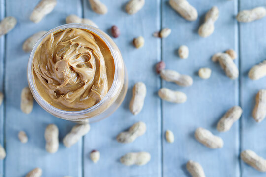 Table top view of swirl creamy peanut butter in a plastic jar container over a blue table with scattered nuts in the shell. Selective focus with blurred background. 