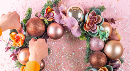 Female hand decorating beautiful unusual Christmas wreath on pink background. flat lay