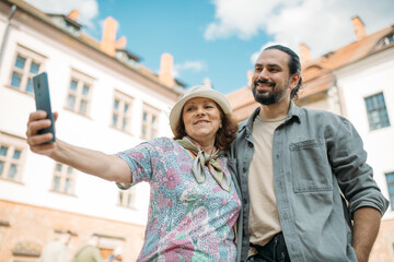 A pensioner and her adult son on a guided tour of an ancient castle. They smile happily and take selfies on their phones. An elderly mother and son travel together, visiting historical sites