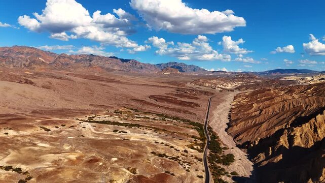Flight over the highway along the dry desert and bare rocks. White fluffy clouds hang in the blue sky above Death Valley, California, United States.