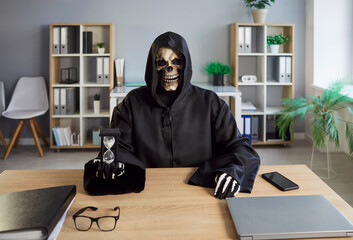 Scary macabre horror Grim Reaper in hooded cloak at office desk with laptop computer and mobile...