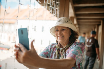 An elderly, contented woman walks with a guide through the park next to an ancient castle. A pensioner inspects the historical architecture and takes pictures on her phone.