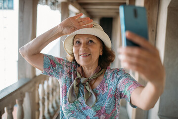 An elderly, contented woman takes a guided tour of the park near the ancient castle. The pensioner examines the historical architecture and takes a selfie with her phone.