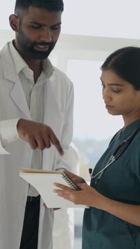 Vertical video indian doctor and young female nurse discussing a patient diagnosis and treatment plan in a bright modern hospital hallway