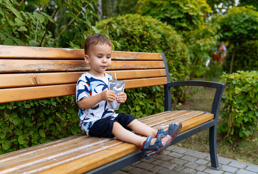 Boy resting with a foil juice pouch. Seated on the wooden bench, the toddler holds a silver drink pouch and enjoys a quiet refreshment among green bushes. - Powered by Adobe