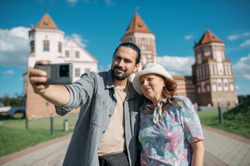A pensioner and her adult son on a guided tour of an ancient castle. They smile happily and take selfies on their phones. An elderly mother and son travel together, visiting historical sites