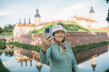 An elderly, contented woman takes a guided tour of the park near the ancient castle. The pensioner examines the historical architecture and takes a selfie with her phone.