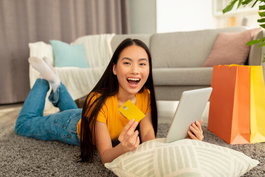 A surprised woman lies on the floor with a tablet and credit card, joyfully reacting to online discounts. Gift bags are nearby, showcasing her excitement for shopping from home.