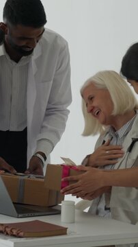 Vertical video diverse medical team surprises senior female physician with birthday gifts in cheerful hospital clinic, smiling and celebrating together