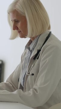 Vertical video mature female physician in lab coat with stethoscope sits at desk, focused on typing and reviewing electronic medical records