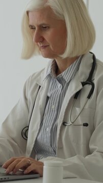 Vertical video experienced female physician wearing a white lab coat and using a stethoscope, seated at a desk while working on a laptop computer in a modern clinic setting, with a white background