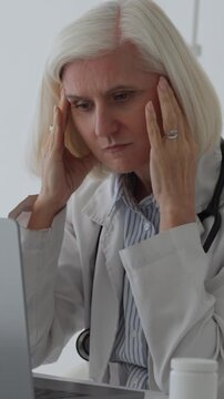 Vertical video middle-aged silver-haired female doctor looking exhausted and stressed, rubbing her temples while working on a laptop in a clinic against a plain white background, burnout