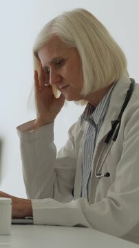 Vertical video elderly female doctor in lab coat sitting at hospital desk, rubbing temples from severe headache and burnout after long shifts