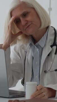 Vertical video anxious senior female physician staring at a container of medication, experiencing anxiety and a migraine while seated at her workstation in a medical facility