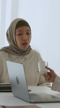Vertical video muslim female doctor in hijab at desk with laptop explaining prescription and handing medicine bottle to patient during consultation