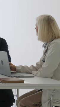 Vertical video mature woman physician in white coat with stethoscope pointing at laptop while discussing diagnosis with male patient in office