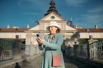 An elderly, contented woman walks with a guide through the park next to an ancient castle. The pensioner examines the historical architecture, looks at a map and reads about the sights on her phone.