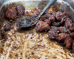 Caramelized Asian-style glazed chicken pieces or wings, sprinkled with sesame seeds, in a metal serving tray with a greasy spoon