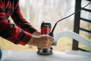 A young man is working with a milling cutter on the veranda of a house. Close-up of hands holding the machine. The carpenter is chamfering plywood using the milling machine.