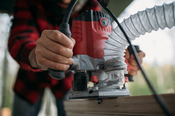 A young man is working with a milling cutter on the veranda of a house. Close-up of hands holding the machine. The carpenter is chamfering plywood using the milling machine.