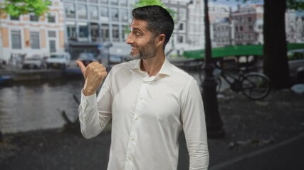 Man bearded wearing white shirt points thumb to canal on street in amsterdam while smiling; confidence welcome.