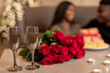 Unrecognizable black couple celebrates Valentine holiday indoors, sitting at a table with wine glasses, a bouquet of red roses, and a gift box nearby, enjoying a romantic moment together.