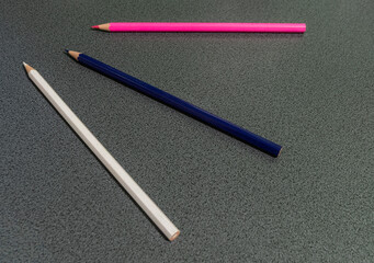 Three colored pencils—pink, blue, and white—arranged diagonally on a gray textured surface. Minimalist composition representing creativity, contrast, and artistic simplicity
