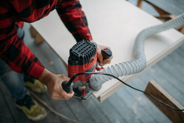 A young man is working with a milling cutter on the veranda of a house. Close-up of hands holding the machine. The carpenter is chamfering plywood using the milling machine.