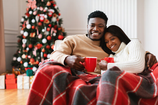 Couple relaxes on sofa covered with a blanket, drinking coffee while watching holiday movies on Christmas Eve. A decorated tree and gifts create a warm, festive atmosphere. - Powered by Adobe