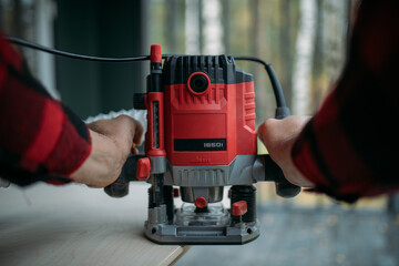 A young man is working with a milling cutter on the veranda of a house. Close-up of hands holding the machine. The carpenter is chamfering plywood using the milling machine.