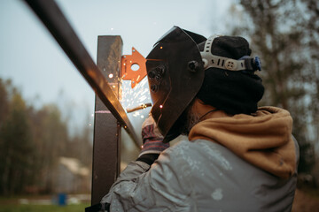 A young man is working as a gas welder on the street. A man wearing a protective mask is welding iron rungs to pillars. The worker, holding a welding machine, sparks fly around him.