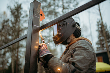 A young man is working as a gas welder on the street. A man wearing a protective mask is welding iron rungs to pillars. The worker, holding a welding machine, sparks fly around him.