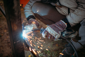A young man is working as a gas welder on the street. A man wearing a protective mask is welding iron rungs to pillars. The worker, holding a welding machine, sparks fly around him.