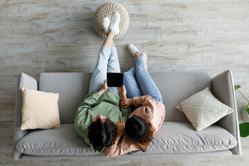 Asian couple sits on a cozy sofa, holding a tablet with a blank screen. They are relaxed and engaged, providing an opportunity for mockup designs or app promotions in a home setting.