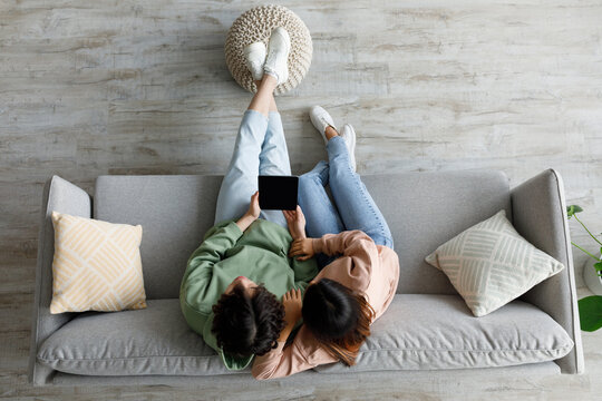 Asian couple sits on a cozy sofa, holding a tablet with a blank screen. They are relaxed and engaged, providing an opportunity for mockup designs or app promotions in a home setting.