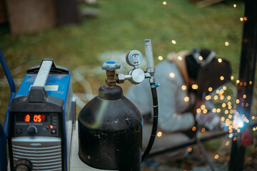 A young man is working as a gas welder on the street. A man wearing a protective mask is welding iron rungs to pillars. The worker, holding a welding machine, sparks fly around him.