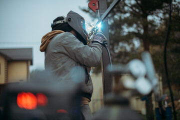 A young man is working as a gas welder on the street. A man wearing a protective mask is welding iron rungs to pillars. The worker, holding a welding machine, sparks fly around him.