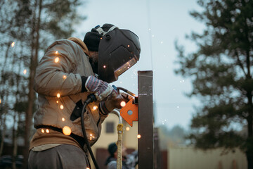 A young man is working as a gas welder on the street. A man wearing a protective mask is welding iron rungs to pillars. The worker, holding a welding machine, sparks fly around him.