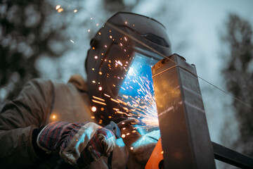 A young man is working as a gas welder on the street. A man wearing a protective mask is welding iron rungs to pillars. The worker, holding a welding machine, sparks fly around him.