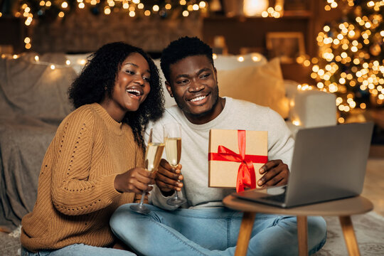 Happy black couple enjoys a festive online gathering during Christmas, holding champagne glasses and sharing a gift while making a video call in their decorated living room filled with lights.