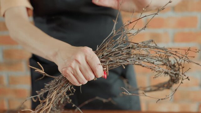 Female florist shaping dry twigs into rustic wreath decoration, hands holding bundle with red nails, creative artisan crafting natural handmade design in workshop with focus on detail and natural