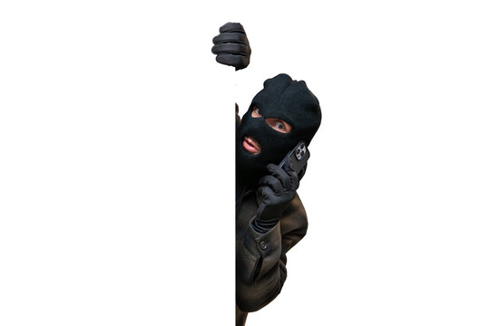 Thief wearing a black balaclava and gloves, peeking from behind a white banner while talking on a smartphone to plan a crime