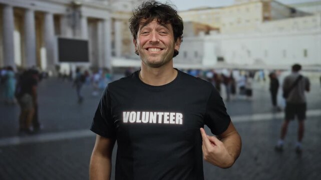Hispanic man in volunteer shirt smiling and pointing at his chest stands in front of a historic monument with columns, creating an inspiring atmosphere on a bustling university campus.