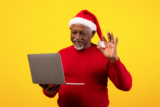 A senior black man is happily making a video call on his laptop while wearing a festive Santa hat. He waves hello as he celebrates Christmas remotely, set against an orange background.