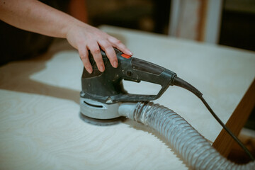 A young woman grinds plywood in a house under construction. Close-up of a woman's hands holding a grinder. A woman works with a manual electric grinder