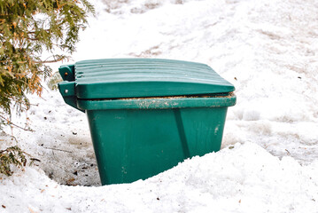 Closeup of green grit container surrounded by snow on city sidewalk, winter preparation and deicing tools for safe walking and driving conditions. Sand and salt mixture storage box on snowy street