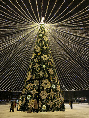 New Year Tree with Glowing Garlands, decorations in town square. People gather around tall coniferous Tree with Stars, Snowflakes in winter. Christmas celebration on a street in Russia. Vertical Photo