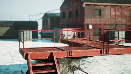 A red platform stands on a stone structure, overlooking a snowy expanse. Nearby, rustic buildings add color to the serene winter scene, illuminated by bright sunlight.